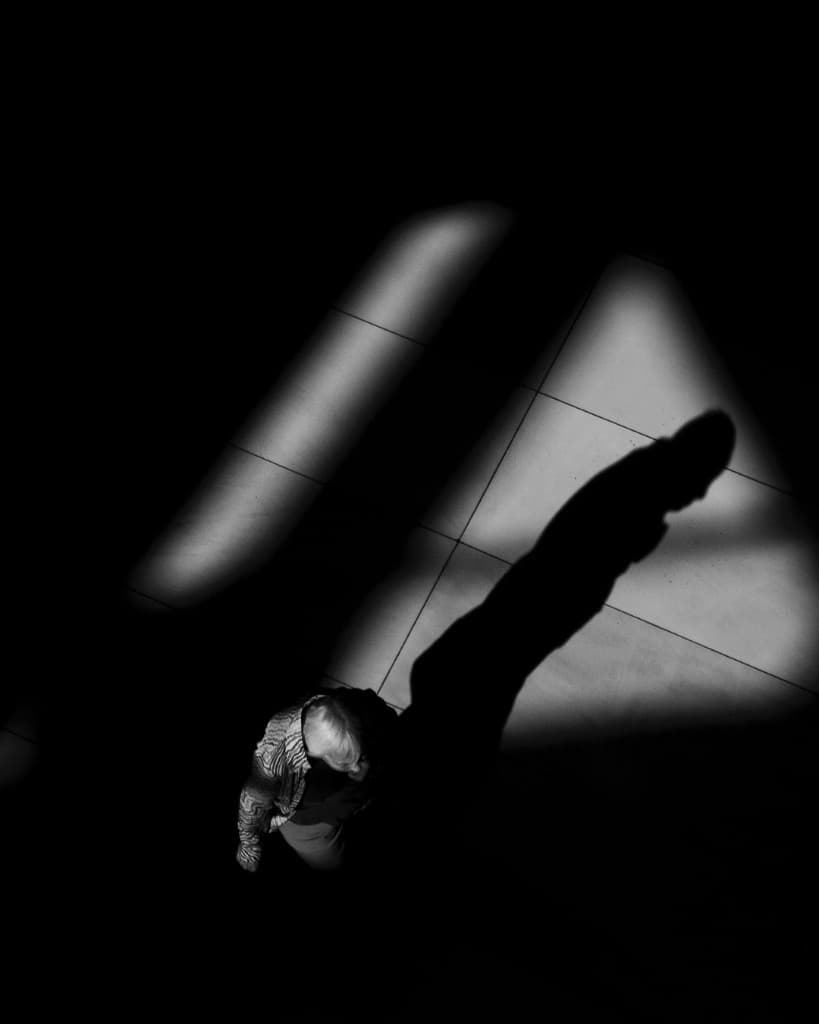 Figura bajo haces de luz proyectando una sombra alargada sobre el piso.