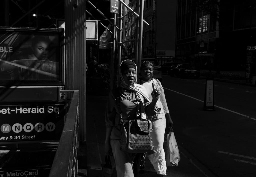 Dos figuras caminan junto a la entrada del metro Herald Square, en luz y sombra.