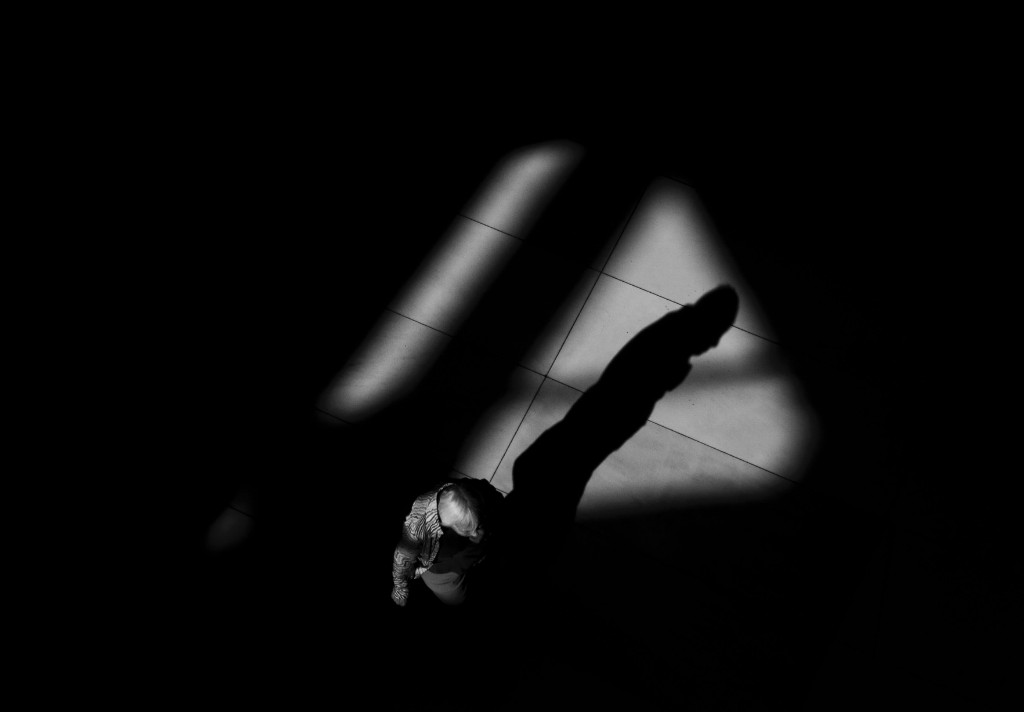 Overhead view of a figure on a tiled floor, crossing a sharp pool of light and shadow.